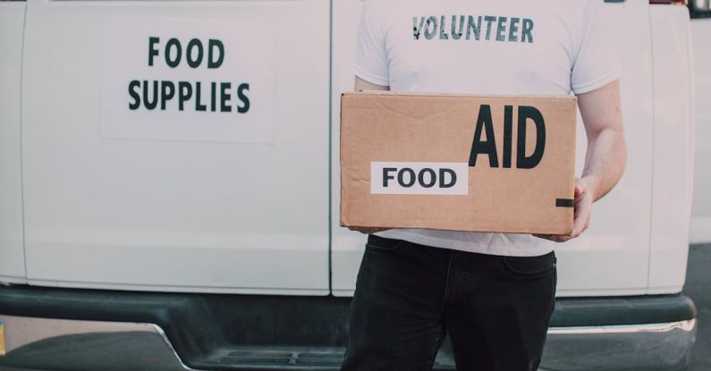 Family Volunteering - Man Wearing White Crew Neck Volunteer T-shirt Holding a Food Labelled Cardboard Box Behind White Van
